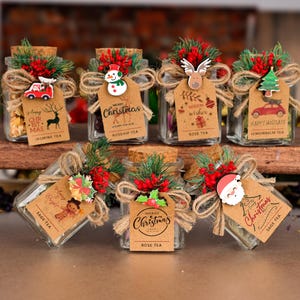 May include: Glass jars filled with tea, decorated for Christmas. Each jar has a cork lid, twine, greenery, and a tag with text like "Merry Christmas" and tea names. Various festive charms are attached.