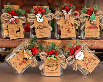 May include: Glass jars filled with tea, decorated for Christmas. Each jar has a cork lid, twine, greenery, and a tag with text like "Merry Christmas" and tea names. Various festive charms are attached.