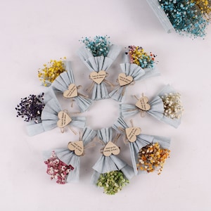 Op de afbeelding: Assortiment van kleine boeketten gedroogde bloemen in verschillende kleuren, verpakt in blauw tissuepapier en vastgebonden met touw. Elk boeket heeft een klein houten hartvormig label met een boodschap.