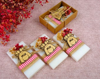 May include: Four white rectangular soaps wrapped in hessian and red and white gingham ribbon. Each soap has a wooden cutout of a girl's dress with the name "Mia" on it.