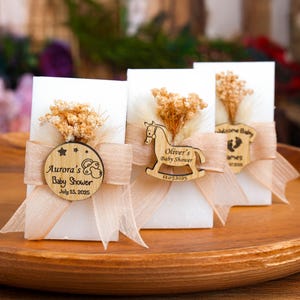 May include: Three white baby shower favors with wooden embellishments and dried floral accents. Each favor is adorned with a light peach ribbon and a wooden tag with the baby's name and shower date. The favors are displayed on a wooden tray.