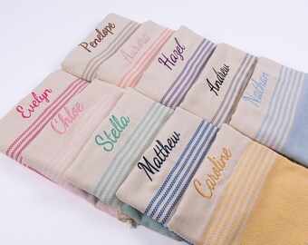 May include: A collection of personalized beige towels with embroidered names in various colors. Each towel features striped accents and a different name, including "Penelope," "Aurora," and "Hazel."