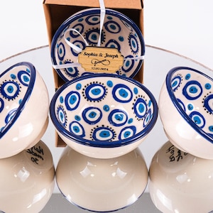 Op de afbeelding: Vier kleine witte keramische kommen met blauwe en witte boze oog-ontwerpen. De kommen zijn in een cirkel op een reflecterend oppervlak gerangschikt. De kommen zijn versierd met een blauwe rand en een blauw en wit boze oog-patroon.