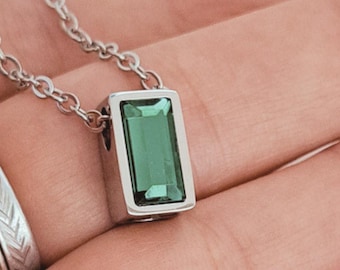 Op de afbeelding: Een zilverkleurige ketting met een rechthoekige hanger met een gefacetteerde smaragdgroene steen, hangend aan een fijne ketting. De hanger zit in een metalen zetting en toont een heldere, levendige groene edelsteen in het midden.