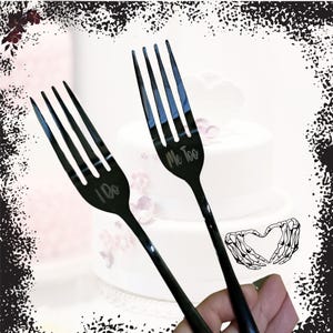 Op de afbeelding: Twee zwarte vorken met de woorden "I Do" en "Me Too" gegraveerd. De vorken staan voor een witte taart met roze bloemen. Een hand houdt de vorken vast.