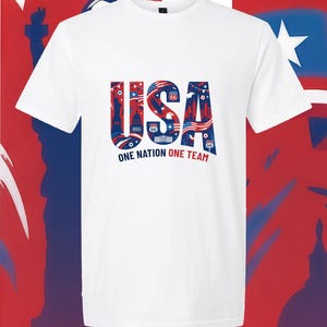 May include: A white short-sleeved t-shirt featuring a large graphic on the chest. The graphic displays the letters USA in blue, filled with red and white patriotic imagery including the Statue of Liberty, an eagle, and Route 66 signs. Below, the text reads ONE NATION ONE TEAM.