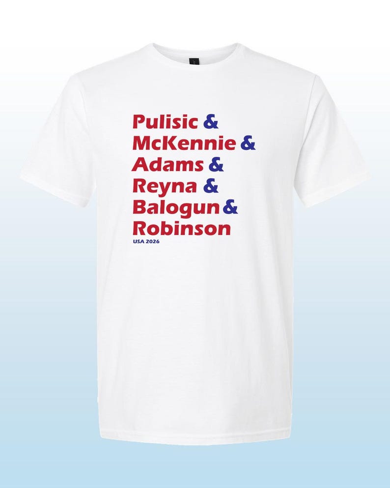 May include: A white short-sleeved t-shirt featuring a list of names in red text followed by blue ampersands. The names listed are Pulisic, McKennie, Adams, Reyna, Balogun, and Robinson. The text USA 2026 is printed in blue at the bottom of the list.