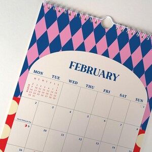 May include: A spiral-bound calendar featuring the month of February. The top portion has a pink and blue diamond pattern. The calendar has a white background with the days of the week and dates. The word "FEBRUARY" is printed in navy blue.