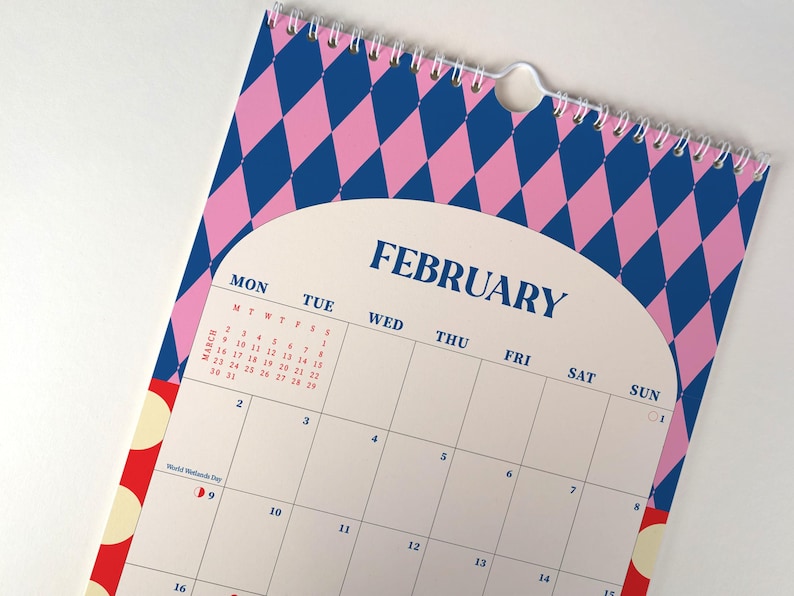 May include: A spiral-bound calendar featuring the month of February. The top portion has a pink and blue diamond pattern. The calendar has a white background with the days of the week and dates. The word "FEBRUARY" is printed in navy blue.