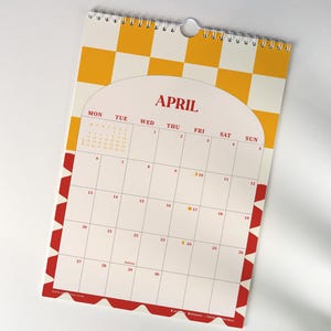 May include: A spiral-bound wall calendar for April, featuring a white background with a yellow and white checkered pattern at the top. The calendar has a red border with a repeating geometric design. The month's name is in red.