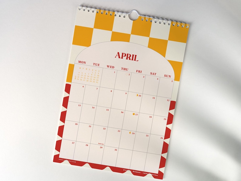 May include: A spiral-bound wall calendar for April, featuring a white background with a yellow and white checkered pattern at the top. The calendar has a red border with a repeating geometric design. The month's name is in red.