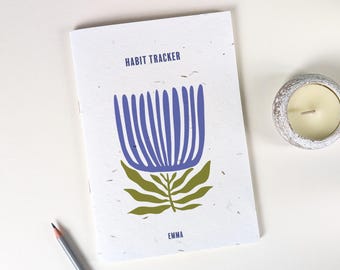 Könnte beinhalten: Ein weißes Notizbuch zur Gewohnheitsverfolgung mit einem blauen Blumendesign und dem Namen "EMMA" unten. Ein Bleistift und ein kleiner Kerzenhalter mit einer brennenden Kerze sind ebenfalls zu sehen.