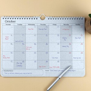 May include: A horizontal wall calendar for October with a speckled light grey background, black wire binding, and a metal hanger. The grid features handwritten notes in blue and red ink, with a monthly focus section and a notes area at the bottom.