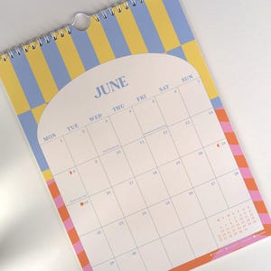 May include: A spiral-bound wall calendar for June, featuring a retro design with a yellow and blue striped background. The calendar has a white interior with dates and days of the week, and a colorful border.
