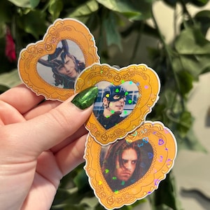 May include: Heart-shaped stickers featuring portraits of characters in ornate gold frames. The frames have a floral design and iridescent glitter. The portraits include a person with horns, a person wearing a mask, and a person with long hair.