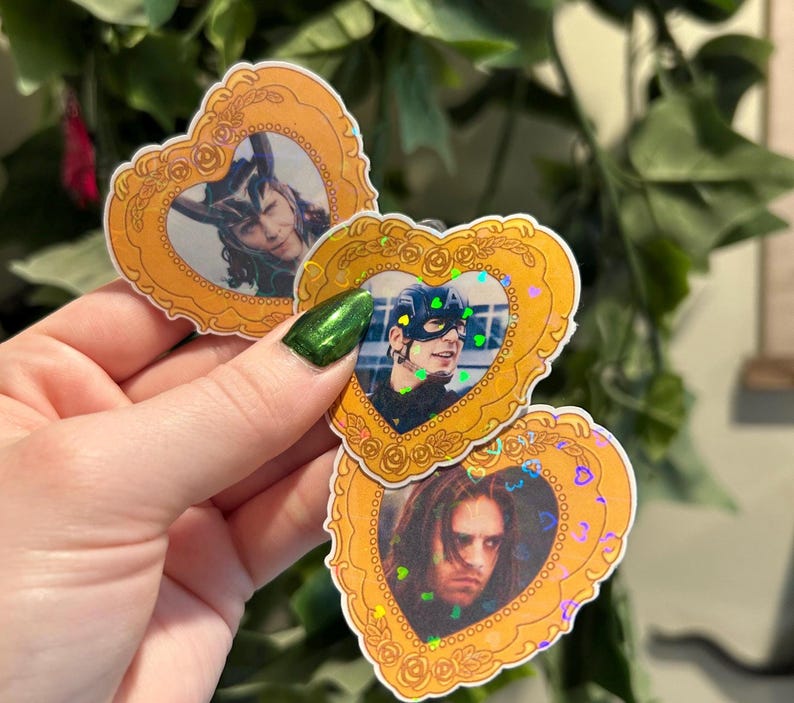 May include: Heart-shaped stickers featuring portraits of characters in ornate gold frames. The frames have a floral design and iridescent glitter. The portraits include a person with horns, a person wearing a mask, and a person with long hair.