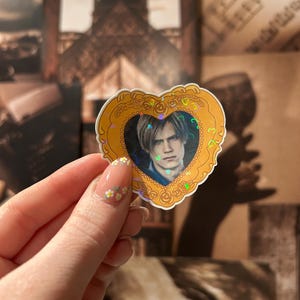 May include: A heart-shaped sticker with a gold border and a portrait of a person with light brown hair. The portrait is surrounded by a holographic effect. The sticker is held up against a background of sepia-toned images.