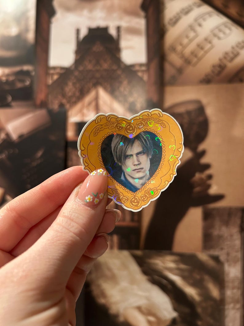 May include: A heart-shaped sticker with a gold border and a portrait of a person with light brown hair. The portrait is surrounded by a holographic effect. The sticker is held up against a background of sepia-toned images.