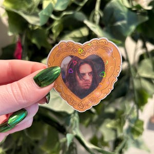 May include: A heart-shaped sticker featuring a portrait of a person with long hair. The portrait is framed by a gold-colored border with floral and heart accents. Small heart-shaped confetti is scattered around the portrait.