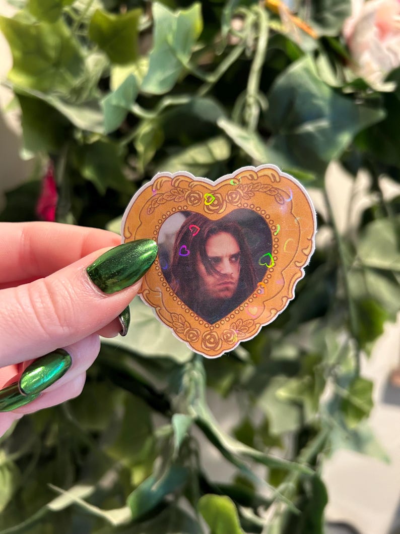 May include: A heart-shaped sticker featuring a portrait of a person with long hair. The portrait is framed by a gold-colored border with floral and heart accents. Small heart-shaped confetti is scattered around the portrait.
