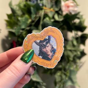 May include: A heart-shaped sticker featuring a portrait of a character with long black hair and a horned helmet. The portrait is set within a gold-colored frame with a decorative border. The sticker has a holographic effect.
