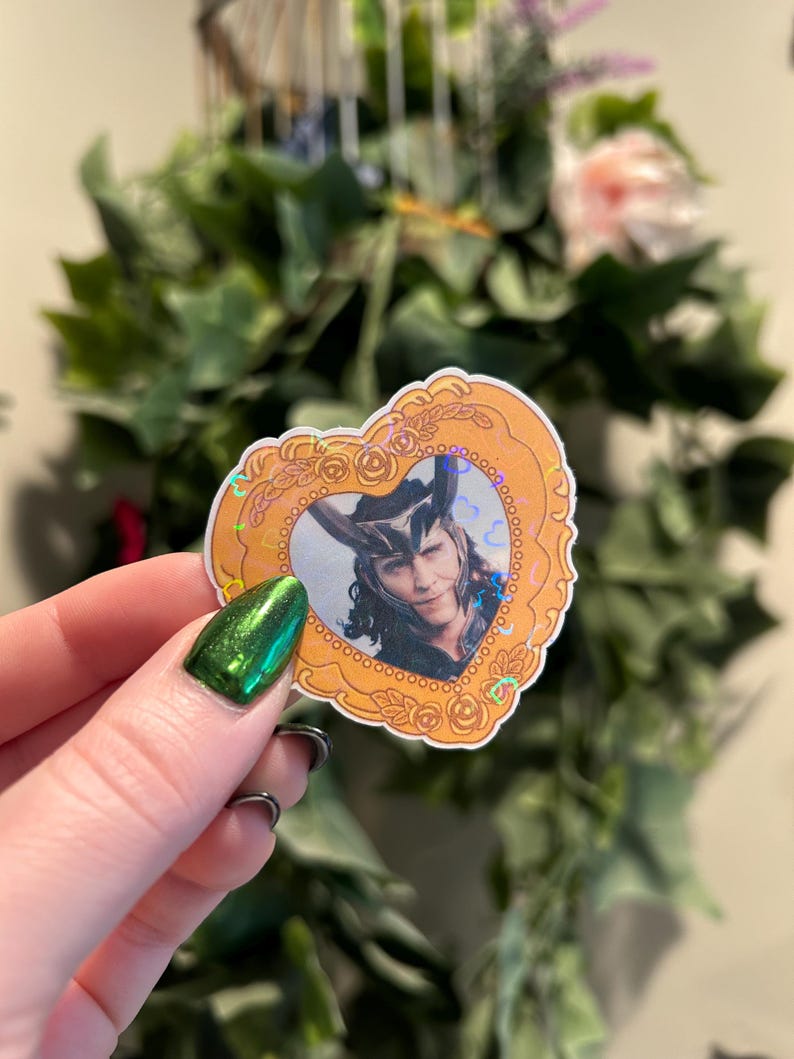 May include: A heart-shaped sticker featuring a portrait of a character with long black hair and a horned helmet. The portrait is set within a gold-colored frame with a decorative border. The sticker has a holographic effect.