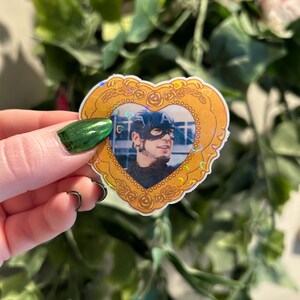 May include: A heart-shaped sticker with a gold border and a photo of a person wearing a helmet. The sticker has a holographic effect and is held up by a hand with green nail polish. The background is blurred with green leaves and a pink flower.