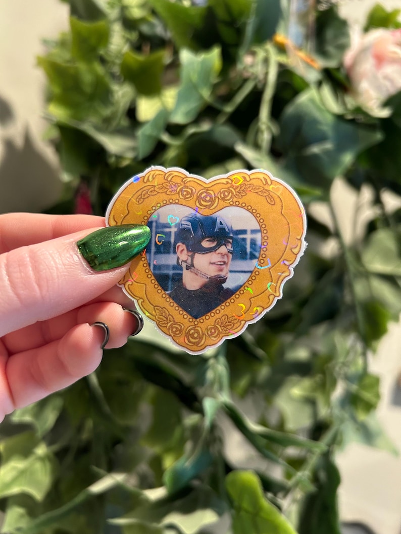 May include: A heart-shaped sticker with a gold border and a photo of a person wearing a helmet. The sticker has a holographic effect and is held up by a hand with green nail polish. The background is blurred with green leaves and a pink flower.