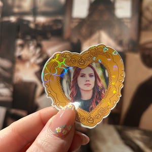 May include: A heart-shaped sticker featuring a portrait of a woman with red hair, framed by a gold border with rose details. The sticker has a holographic effect, creating a rainbow sheen. The background is a blurred image.