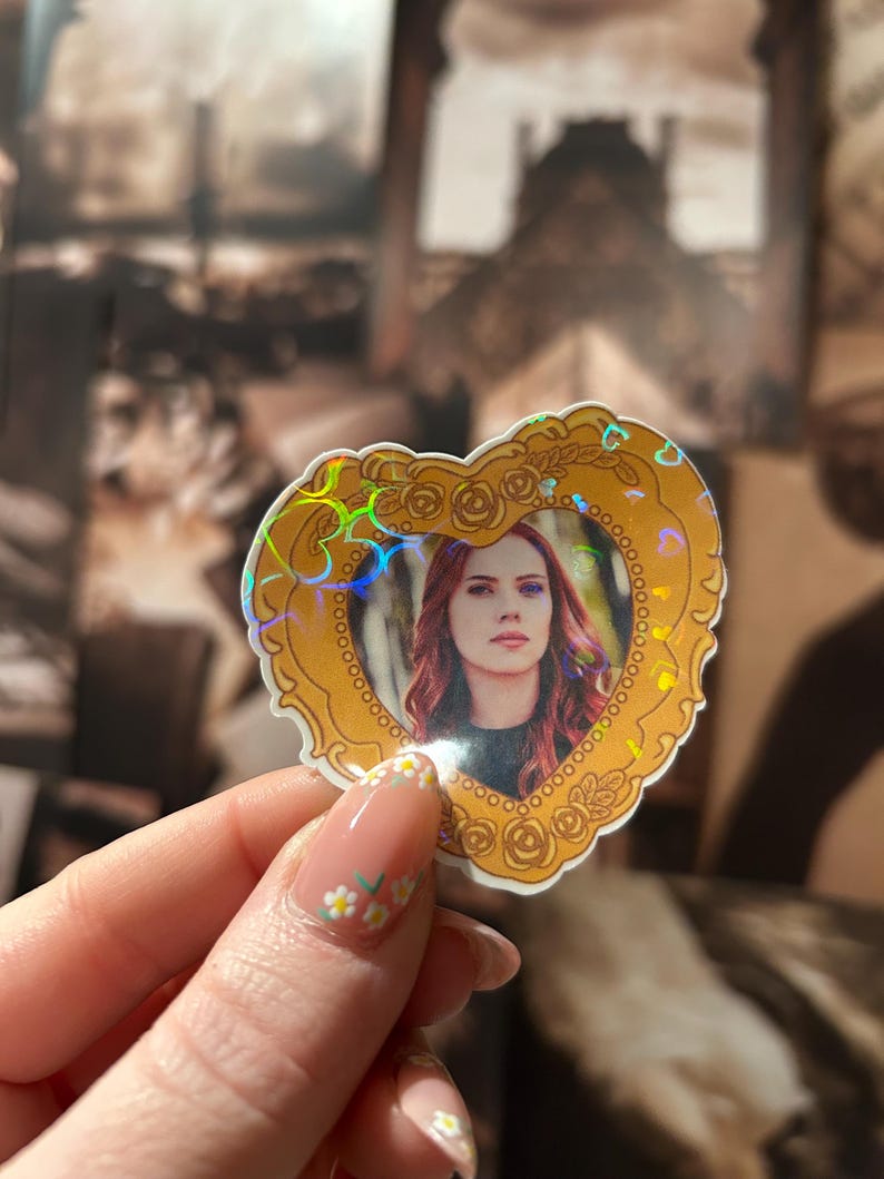 May include: A heart-shaped sticker featuring a portrait of a woman with red hair, framed by a gold border with rose details. The sticker has a holographic effect, creating a rainbow sheen. The background is a blurred image.