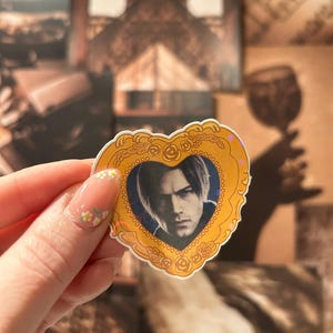 May include: A heart-shaped sticker featuring a portrait of a person with dark hair, framed by a gold-colored border with rose details. The sticker has a blue background and is held up against a backdrop of vintage-style images.