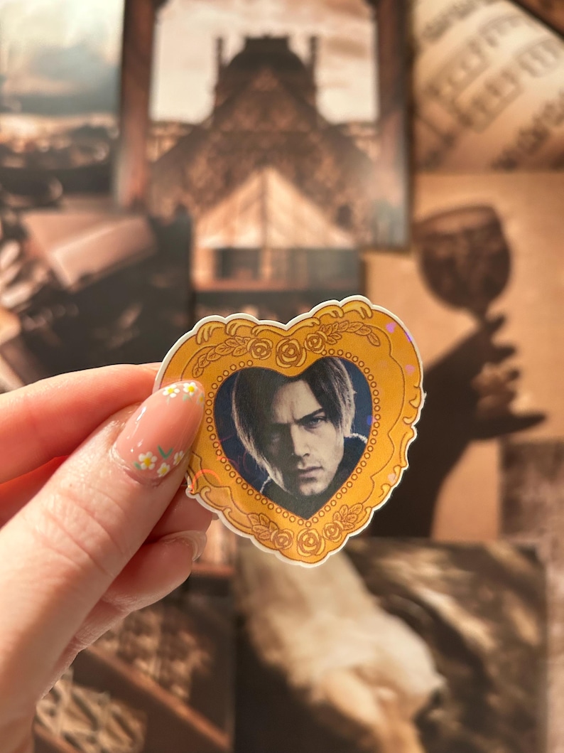 May include: A heart-shaped sticker featuring a portrait of a person with dark hair, framed by a gold-colored border with rose details. The sticker has a blue background and is held up against a backdrop of vintage-style images.