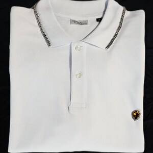 Puede incluir: Camiseta polo blanca con un detalle de cadena plateada en el cuello y una pequeña gema amarilla en el pecho izquierdo.