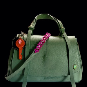 Puede incluir: Un bolso de mano de cuero verde con una correa de joyas rosas y un llavero con el texto "Genuine Leather Made in Italy".