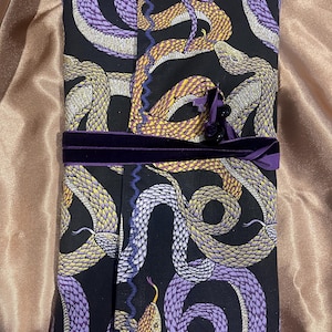 May include: A rectangular fabric wrap featuring a black background with a repeating pattern of snakes in shades of purple, yellow, and metallic gold. The wrap is secured with a dark purple velvet ribbon tied around the centre with small dark beads.