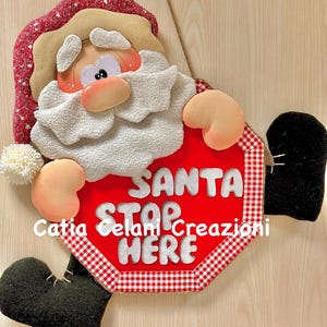 May include: A whimsical Christmas decoration featuring a plush Santa Claus figure holding a red stop sign. The sign reads "Santa Stop Here" in white letters. The Santa figure has a white beard, rosy cheeks, and a red and white hat.