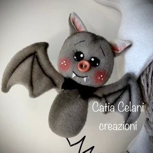 K&ouml;nnte beinhalten: Eine graue Pl&uuml;schfledermaus mit einer rosa Nase und schwarzen Augen. Die Fledermaus h&auml;ngt an einer Feder und hat eine schwarze Schleife. Der Text "Catia Celani creazioni" ist im Bild sichtbar.