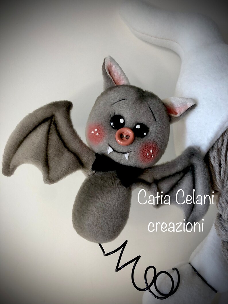K&ouml;nnte beinhalten: Eine graue Pl&uuml;schfledermaus mit einer rosa Nase und schwarzen Augen. Die Fledermaus h&auml;ngt an einer Feder und hat eine schwarze Schleife. Der Text "Catia Celani creazioni" ist im Bild sichtbar.