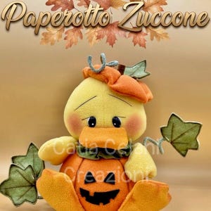 Puede incluir: Un patito de peluche amarillo sentado dentro de una calabaza naranja con una cara de linterna tallada. El pato lleva un pequeño sombrero de calabaza naranja y está rodeado de hojas de tela verde. El texto Paperotto Zuccone aparece arriba.