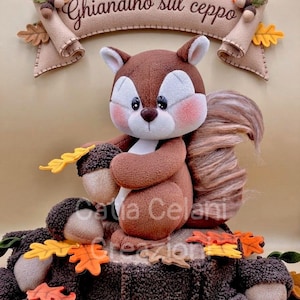 May include: A felt squirrel figurine sitting on a textured brown tree stump, holding an acorn. The squirrel has a fluffy tail and pink cheeks. Above it hangs a beige felt banner with the text Ghiandino sul ceppo, decorated with felt oak leaves and acorns.