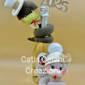 May include: A bottle of sparkling wine adorned with two plush snake figures. One dark gray snake wears a white top hat and gold bow tie, while a light gray snake wears a white pom-pom hat and gold bow. A glittery 2025 topper sits behind the bottle.