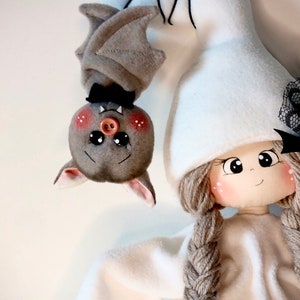 K&ouml;nnte beinhalten: Eine wei&szlig;e Geisterpuppe mit braunen Garnz&ouml;pfen und einer schwarzen Schleife. Die Puppe tr&auml;gt einen wei&szlig;en Hut und hat eine schwarze Knopfnase. Eine graue Fledermaus mit rosa Wangen und einer schwarzen Fliege h&auml;ngt kopf&uuml;ber &uuml;ber der Puppe. Der Text "Catia Celani creazioni" steht &uuml;ber der Puppe.