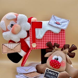 May include: A decorative Santa Claus and reindeer figurine set featuring a red and white chequered mailbox with a white snow-like top. The mailbox has a small wooden tag and a sign that reads Santa's Mailbox, accompanied by several small felt envelopes.