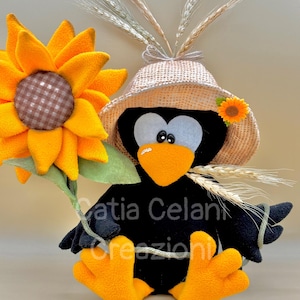 May include: A black crow wearing a straw hat and holding a yellow sunflower. The crow has orange feet and a black beak. The crow is sitting on a light brown surface.