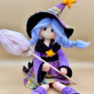 May include: A fabric doll dressed as a witch, featuring light blue yarn hair in braids, a purple and yellow plaid hat with a yellow star, a matching plaid dress with a black collar, a yellow star accent, and black pointed shoes, holding a broom.