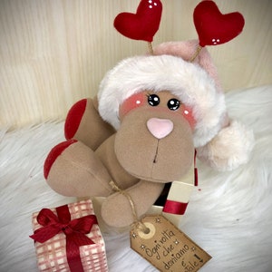 May include: A plush reindeer wearing a Santa hat with red hearts for antlers, sitting in a gift box with a red bow. The box has a tag that reads "Ogni volta che doniamo è Natale!"
