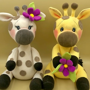 Può includere: Due giocattoli di peluche a forma di giraffa, uno con macchie marroni e un fiore viola, l'altro con macchie gialle e un fiore viola.