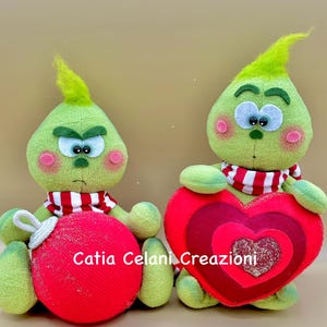 May include: Two green plush Grinch-like figures, one holding a red heart and the other holding a red Christmas ornament. The figures have red cheeks, black eyes, and a red and white striped scarf. The text "Catia Celani Creazioni" is visible on the ornament.