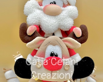May include: A plush Santa Claus doll riding a plush reindeer with a white and brown striped rope. The reindeer has a red nose and black eyes. The Santa doll has a white beard and a red hat. The text "Catia Celani Creazioni" is visible on the reindeer.