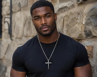 May include: A silver-toned metal cross pendant hangs from a matching silver-toned chain. The cross features a textured, nail-like design with a central horizontal bar. The pendant is worn against a plain, solid black short-sleeved crew neck t-shirt.